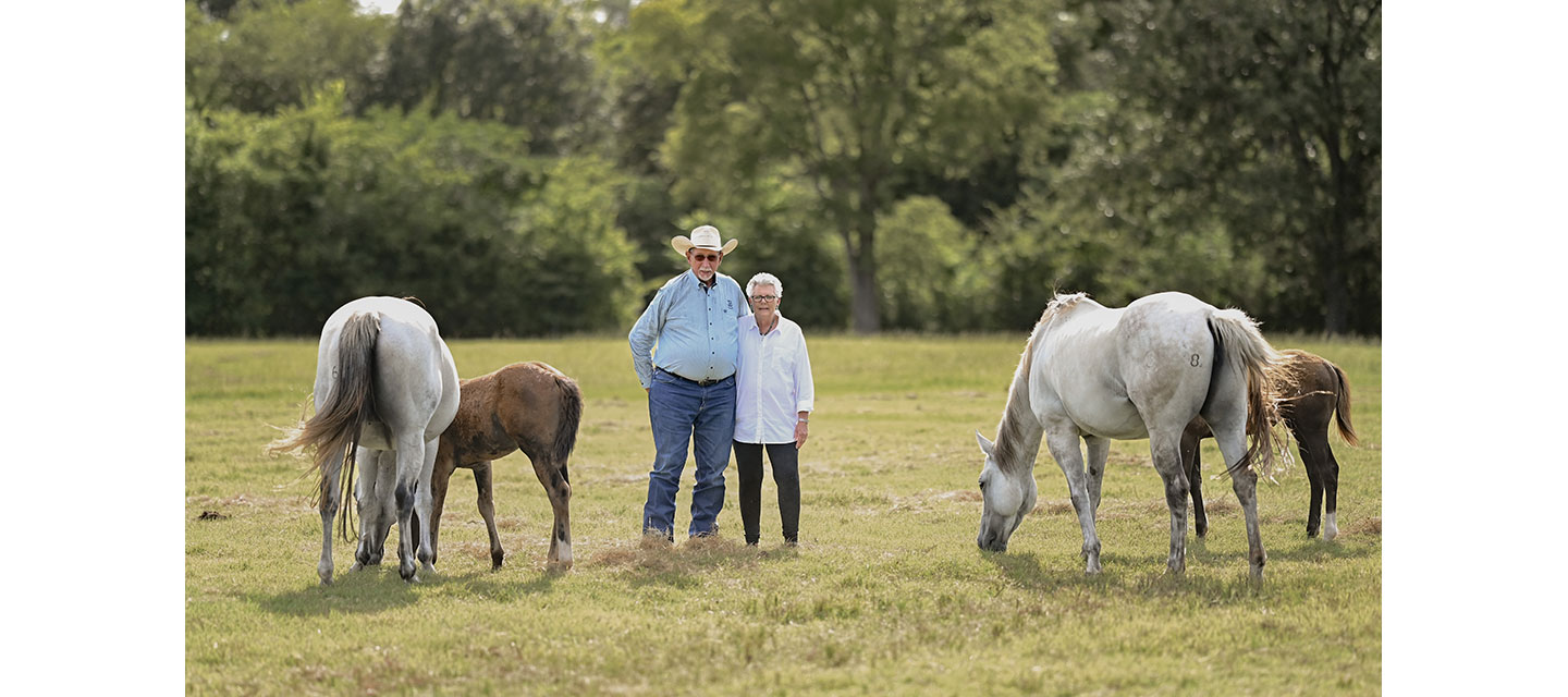An older couple stands close together in the center of a sunlit pasture
